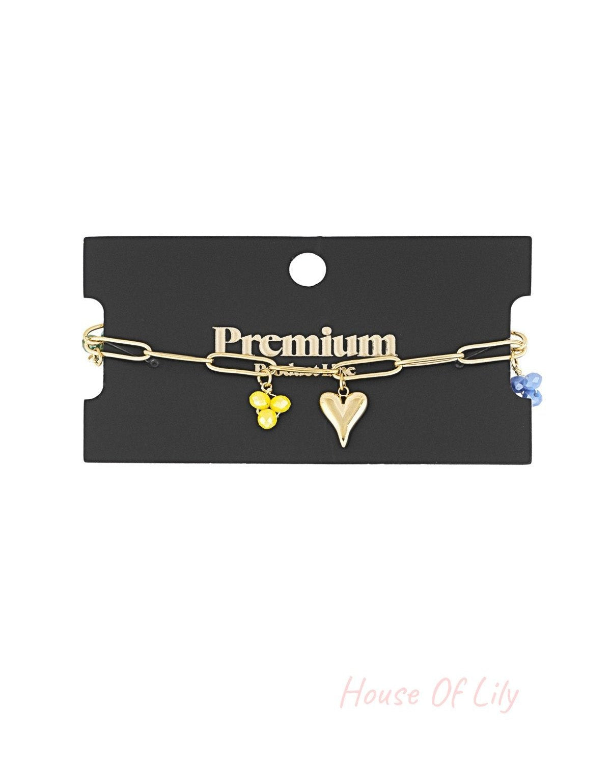 Gold charm bracelet displayed on premium black jewellery card showing yellow and blue bead charms with heart charm