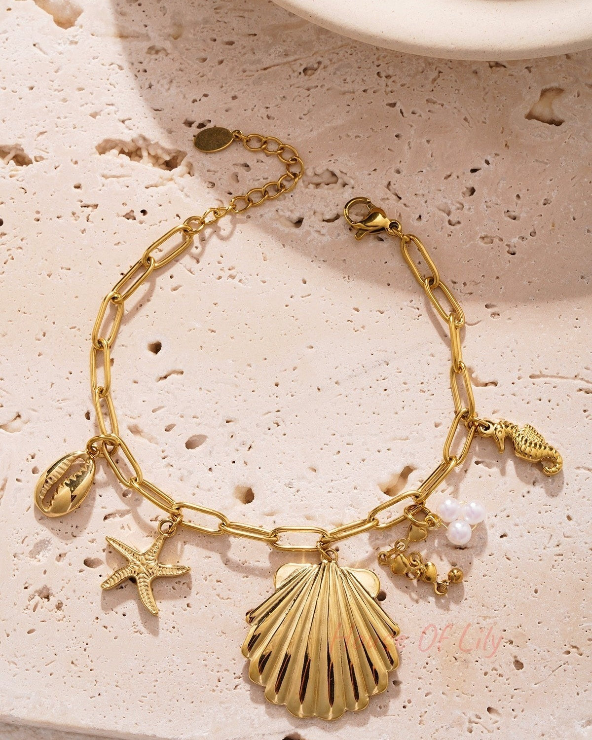 Golden shell charm bracelet placed on textured beige background showcasing lobster clasp and delicate charms
