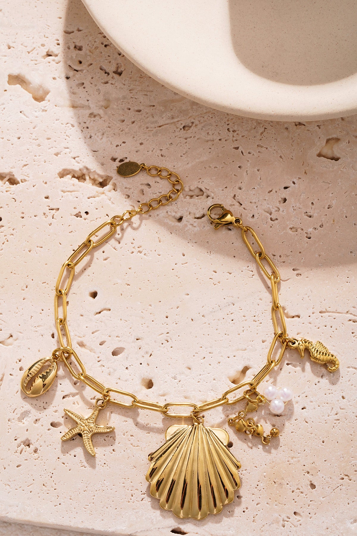 Golden shell charm bracelet placed on textured beige background showcasing lobster clasp and delicate charms
