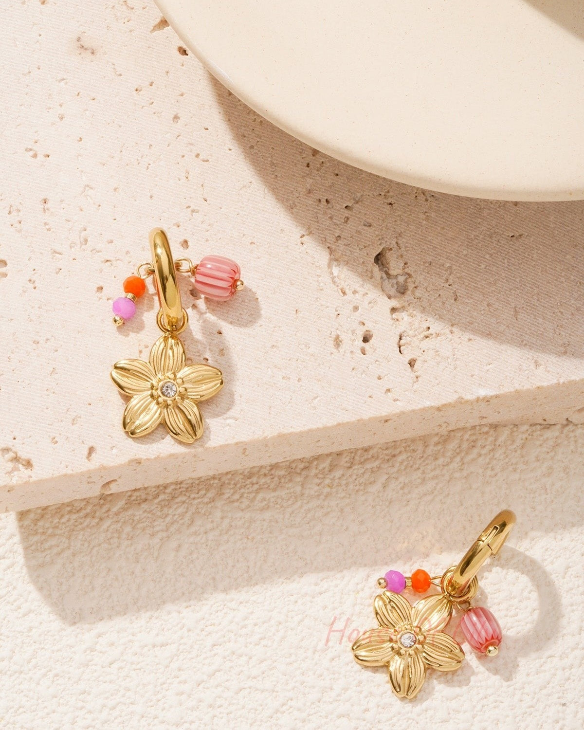 Gold-tone floral charm hoop earrings with colourful beads styled on a textured light stone surface near a cream ceramic object