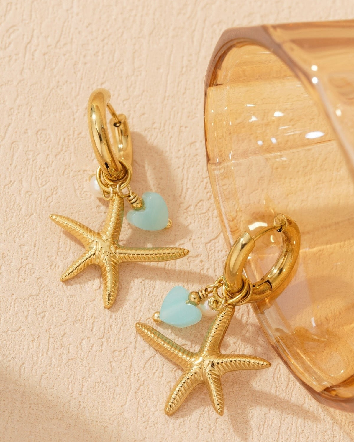 Side view of gold starfish charm hoop earrings leaning against a translucent amber-colored glass