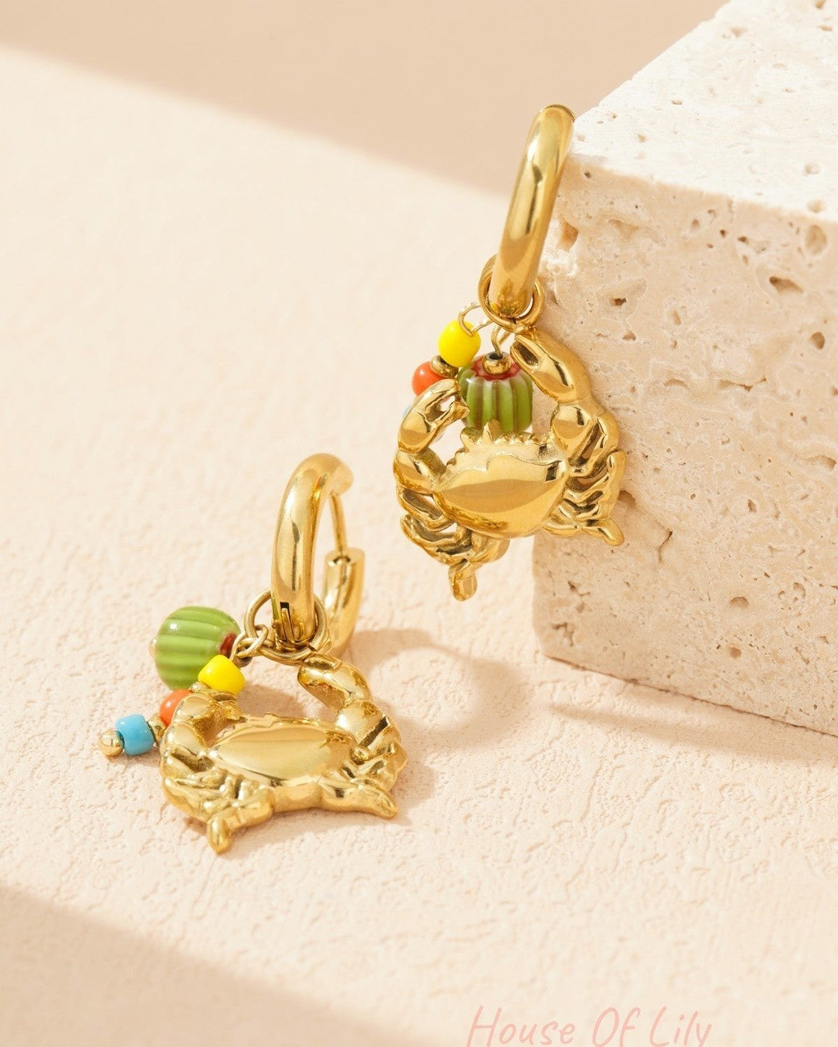Gold-tone crab charm earrings with colourful beads resting against a textured stone surface
