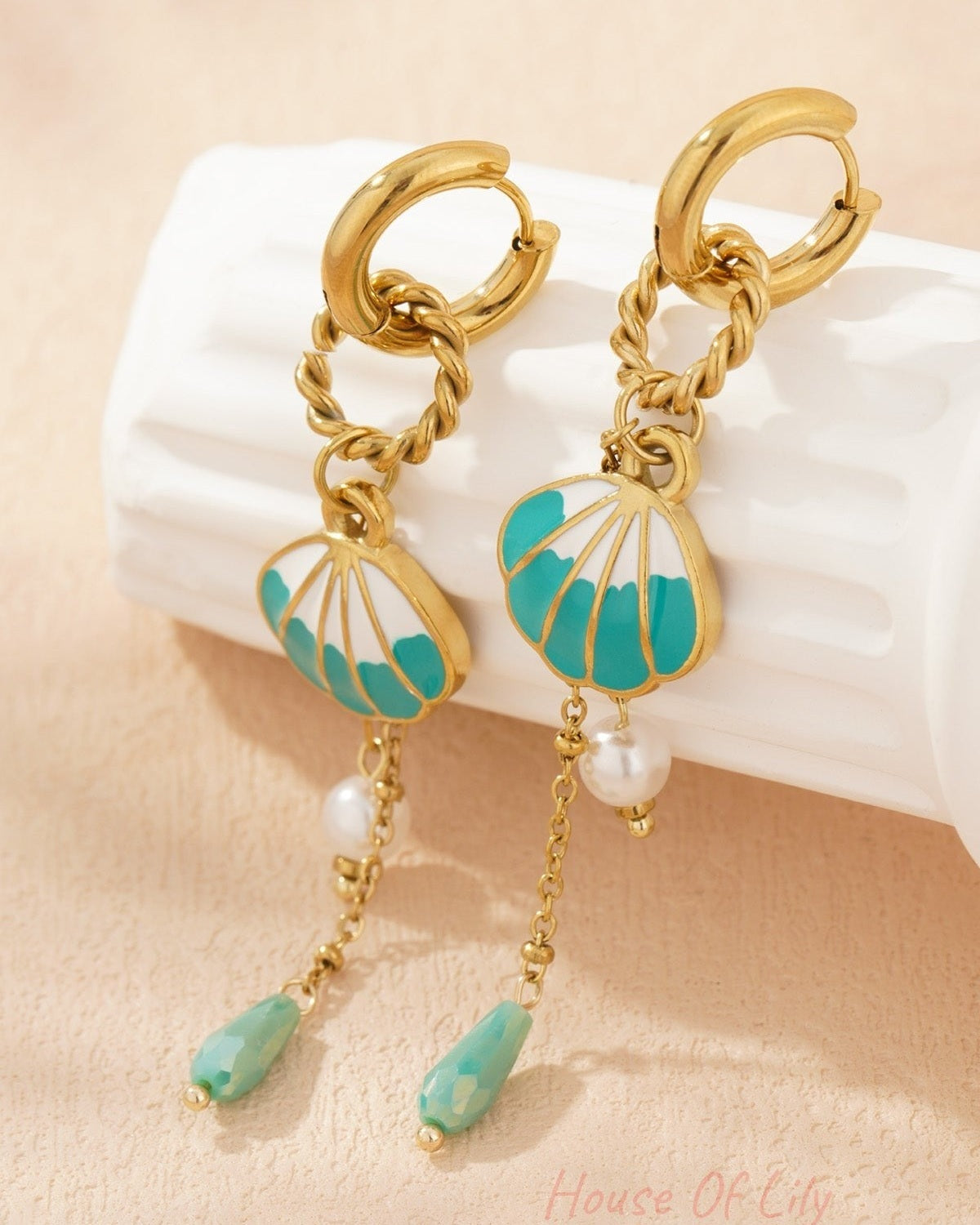 Shell charm drop earrings resting on textured cream background highlighting gold-tone shine, turquoise enamel shell and pearl bead details