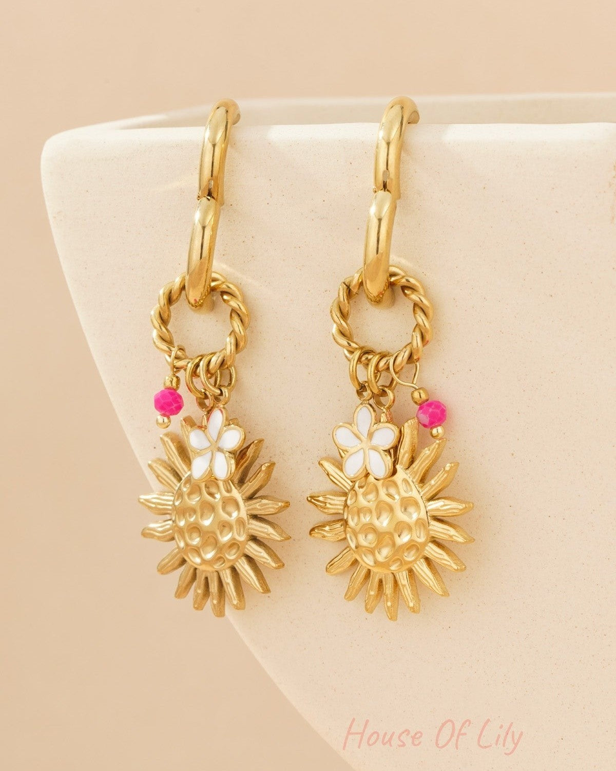 Gold-tone sunshine charm drop earrings hanging on cream-coloured pottery, showing textured sun designs with enamel flowers and pink beads.
