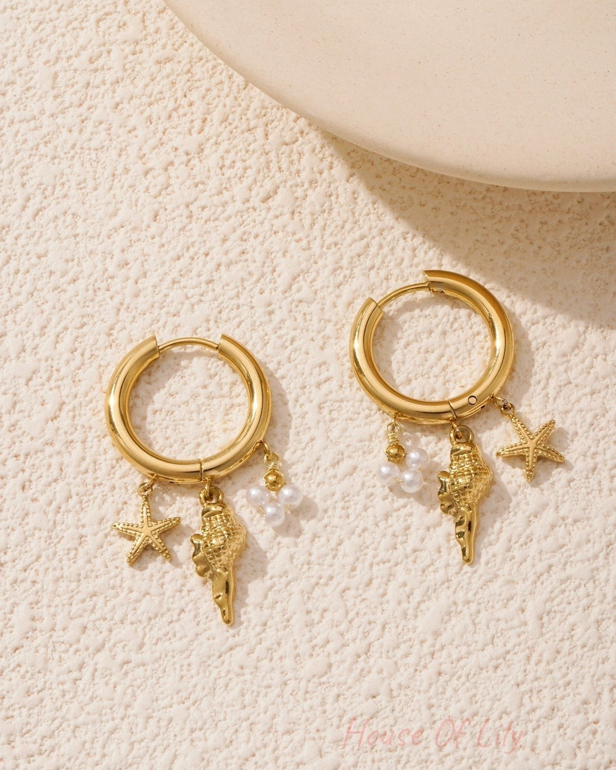 Gold charm hoop earrings placed on textured cream surface with soft lighting