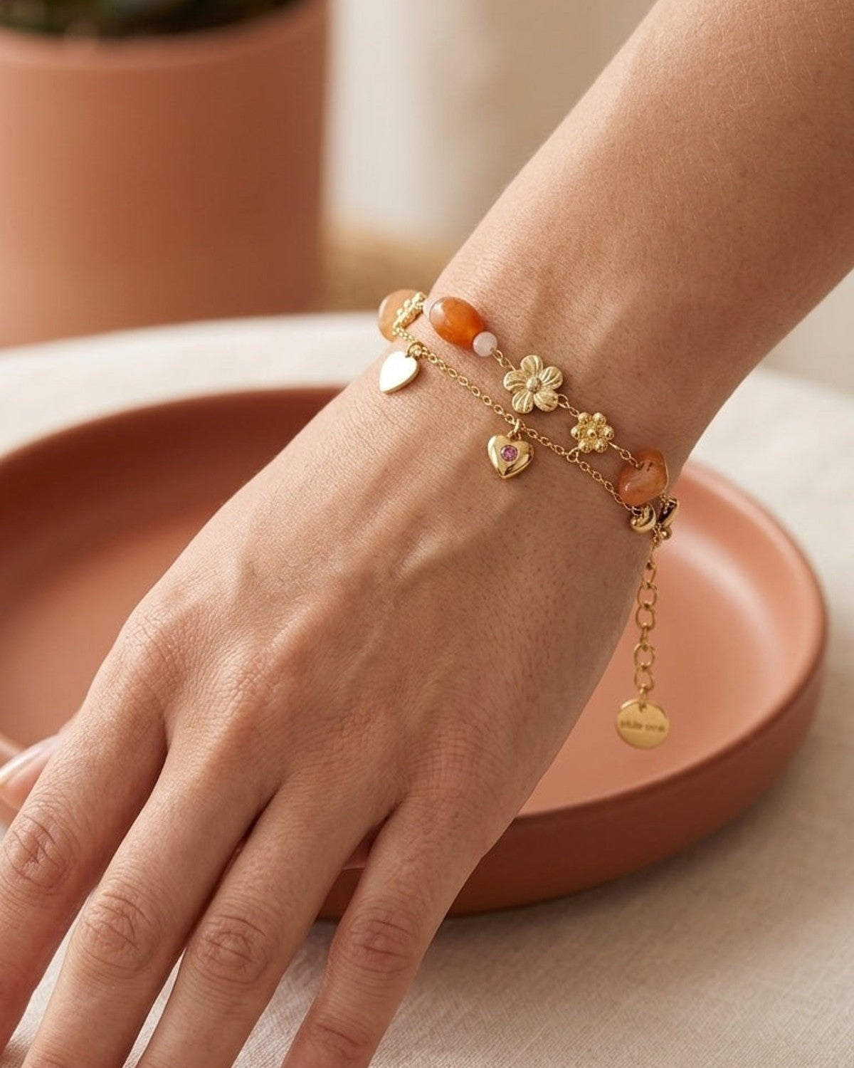 Gold charm bracelet stack with orange gemstones, flower charms, and a heart pendant with a small pink stone.