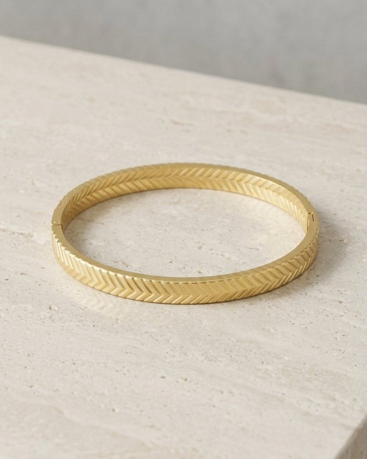 Gold-tone hinged bangle bracelet with a textured herringbone chevron pattern, resting on a beige stone surface.