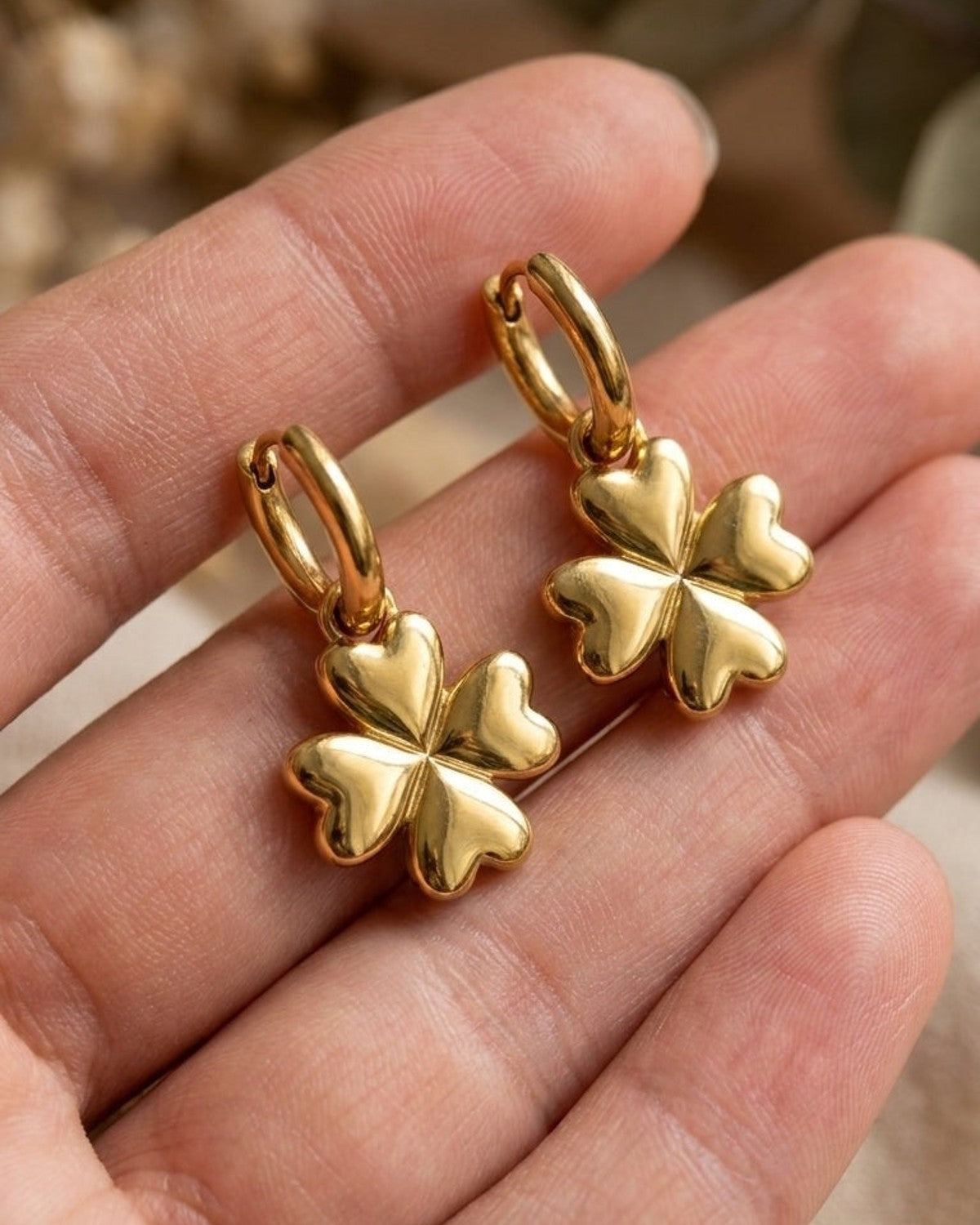 Gold metal drop earrings with small hoop and puffy four-leaf clover charms resting on an open palm.