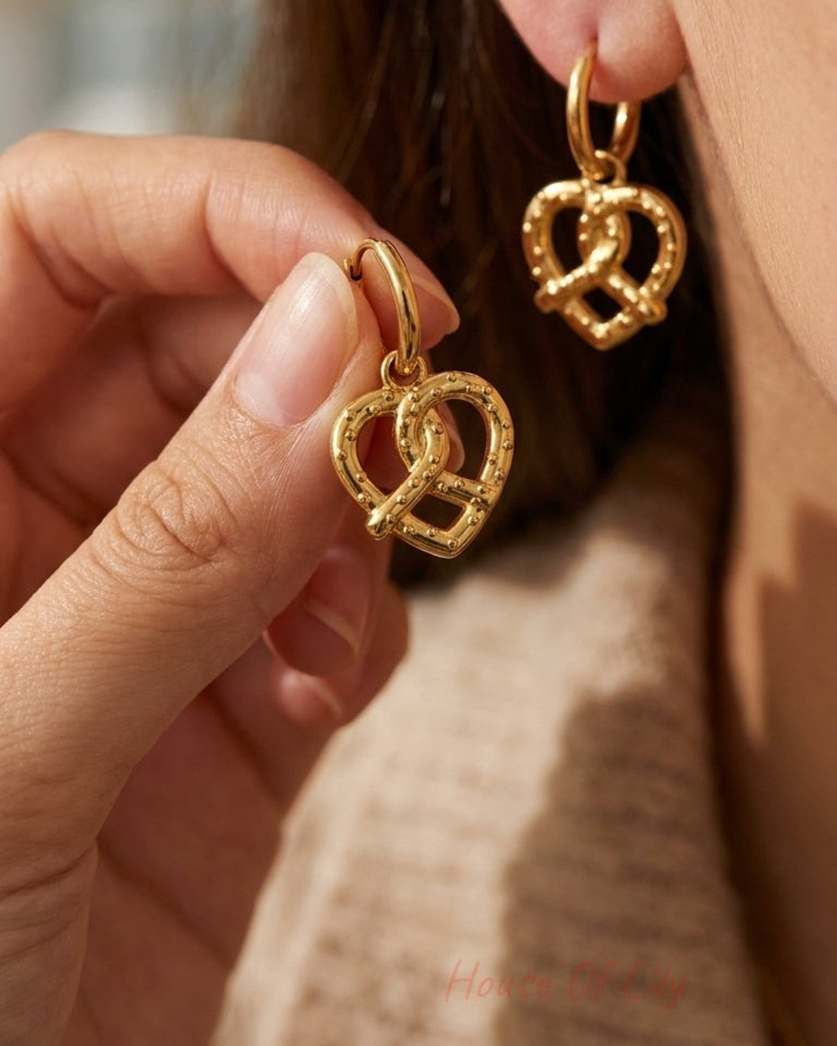 Gold-tone drop earrings featuring a beaded pretzel-shaped charm, worn on an earlobe and held in a hand.