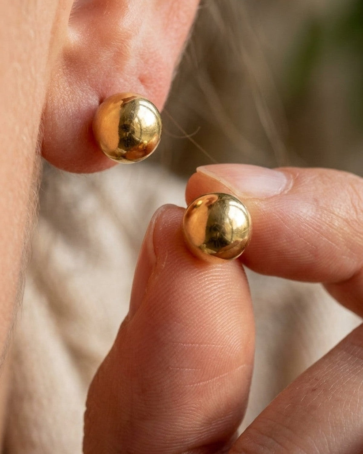 Chic Steel Geometric Studs: polished gold-colored spherical ball stud earrings, one worn on an earlobe and one held in fingers.
