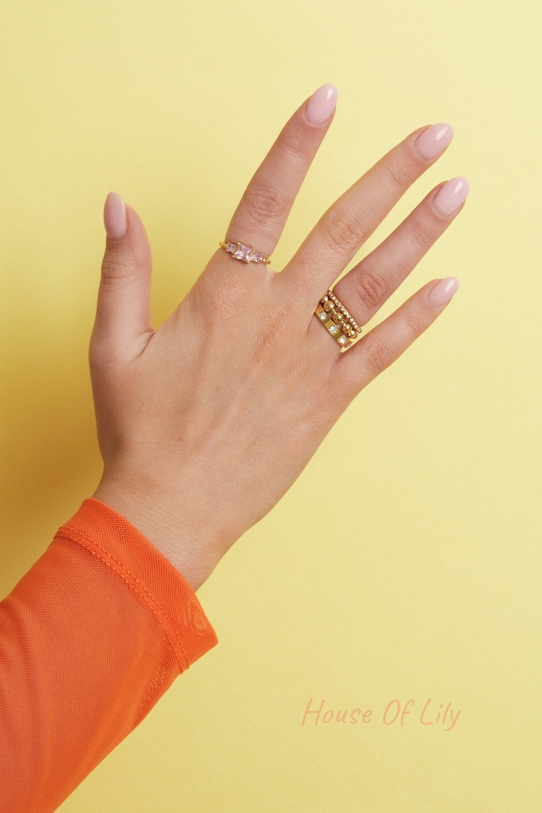 Hand adorned with various gold rings showing different gemstone designs, styled with sheer orange sleeve and yellow background