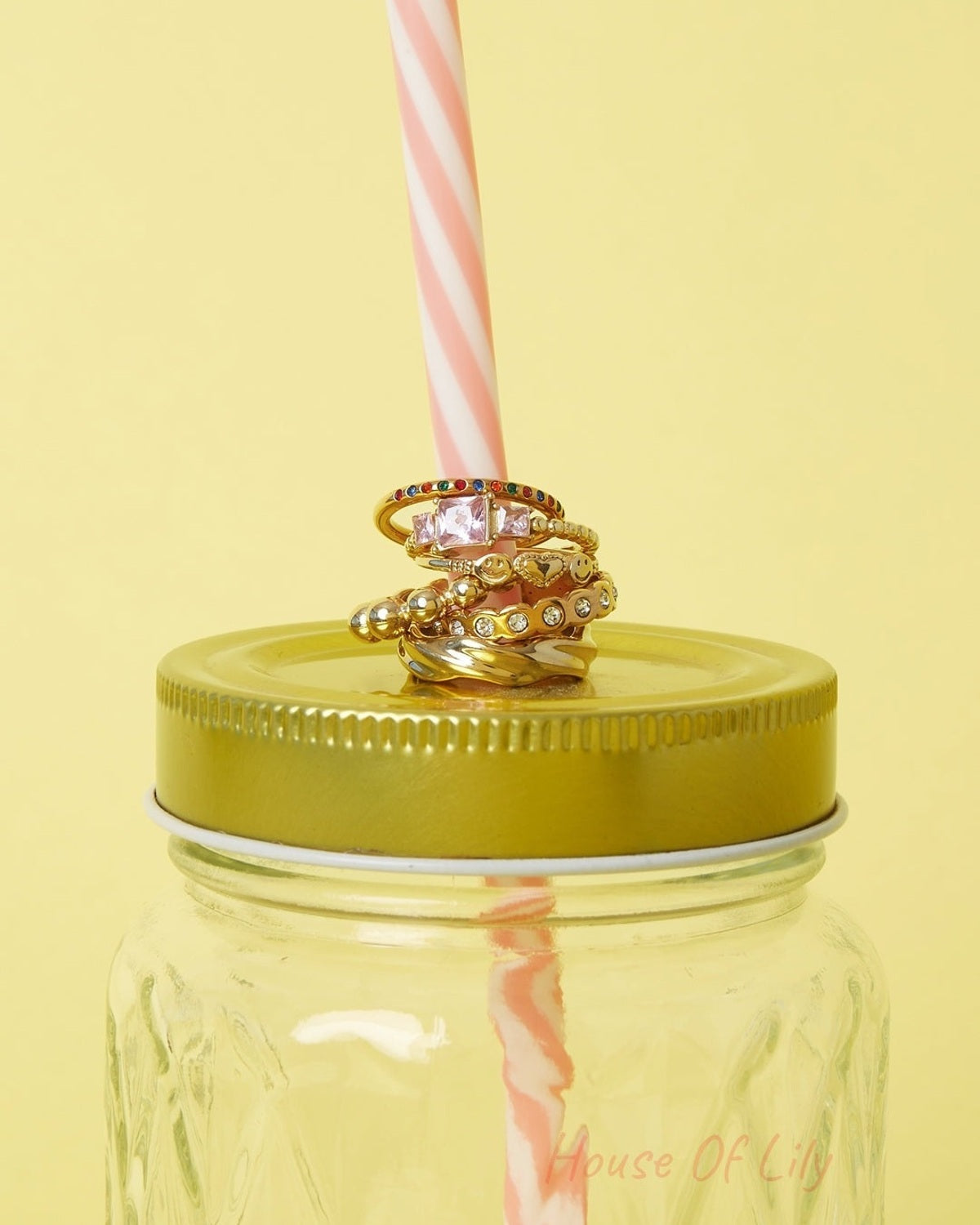 Stack of multiple gold-tone rings, including smiley face and heart designs, displayed on a pink and white striped straw inserted in a yellow-lidded glass jar with a yellow background