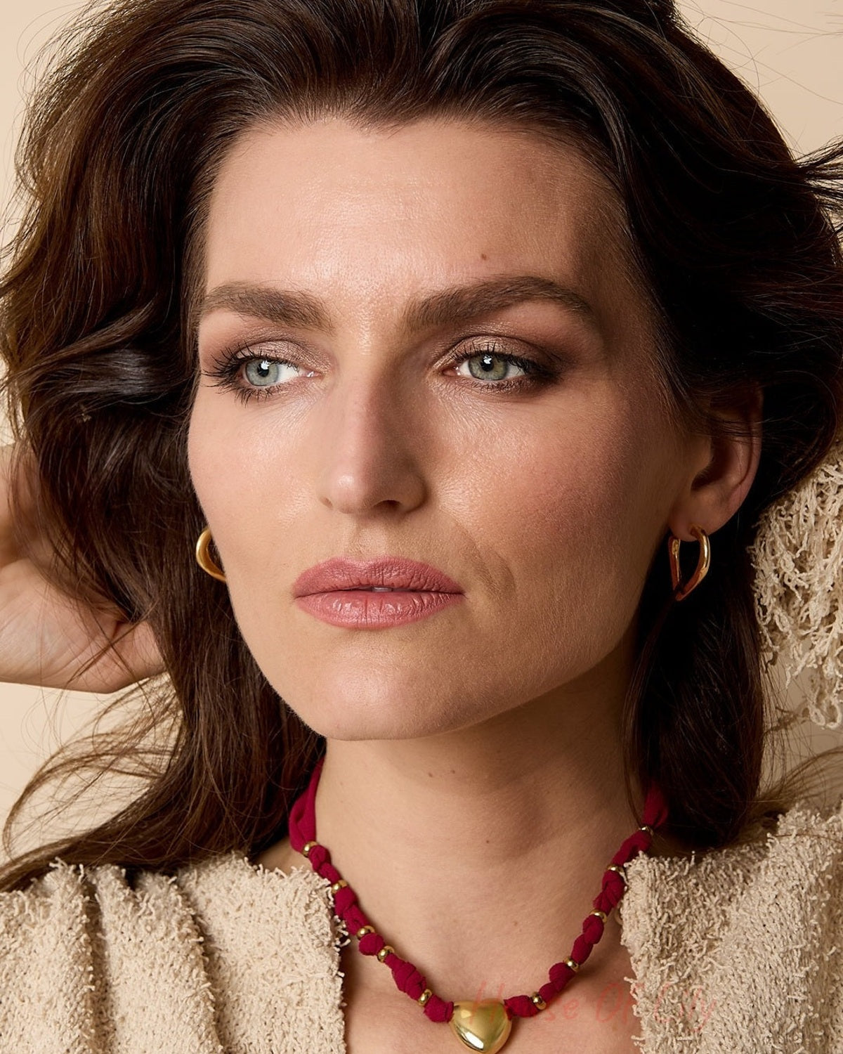 Woman wearing a gold heart pendant necklace with burgundy braided fabric against a beige background