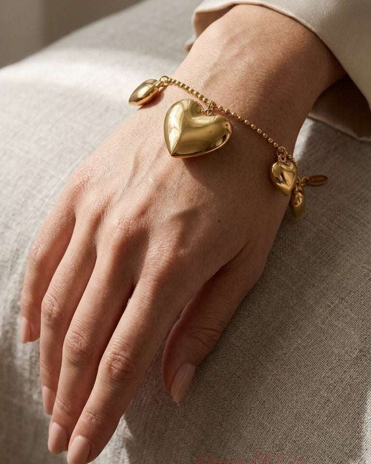 Gold-toned charm bracelet with polished, puffed heart charms on a delicate ball chain, worn on a wrist.