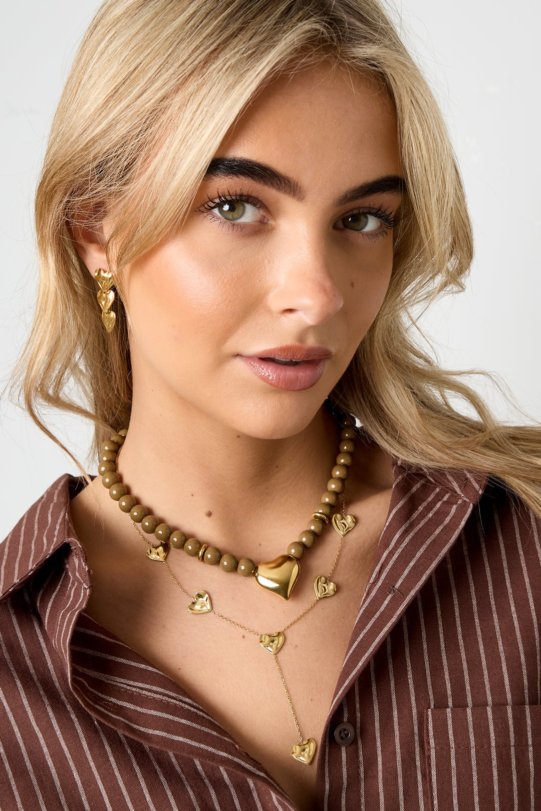 Side angle of model wearing olive bead necklace and layered necklaces with heart pendants, styled with a brown striped top