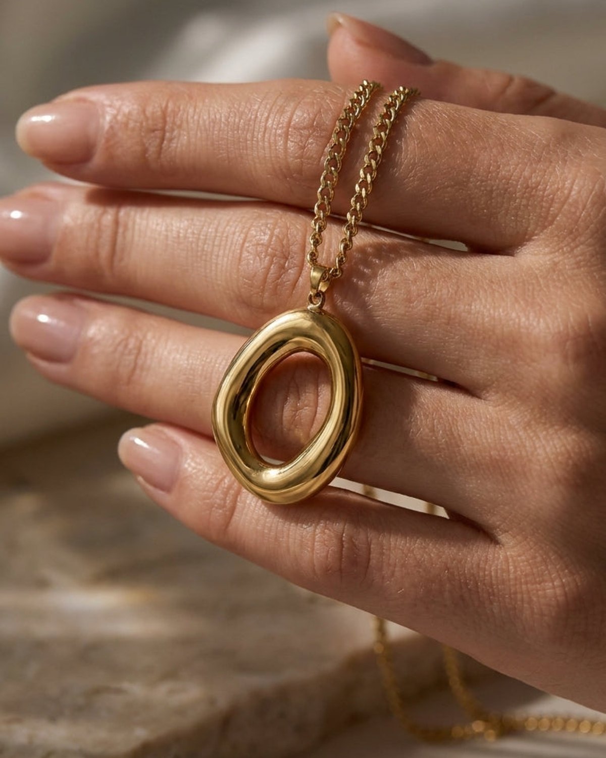Gold-finished oval pendant necklace held in hand, featuring a chunky, organic-shaped hollow oval and matching chain.