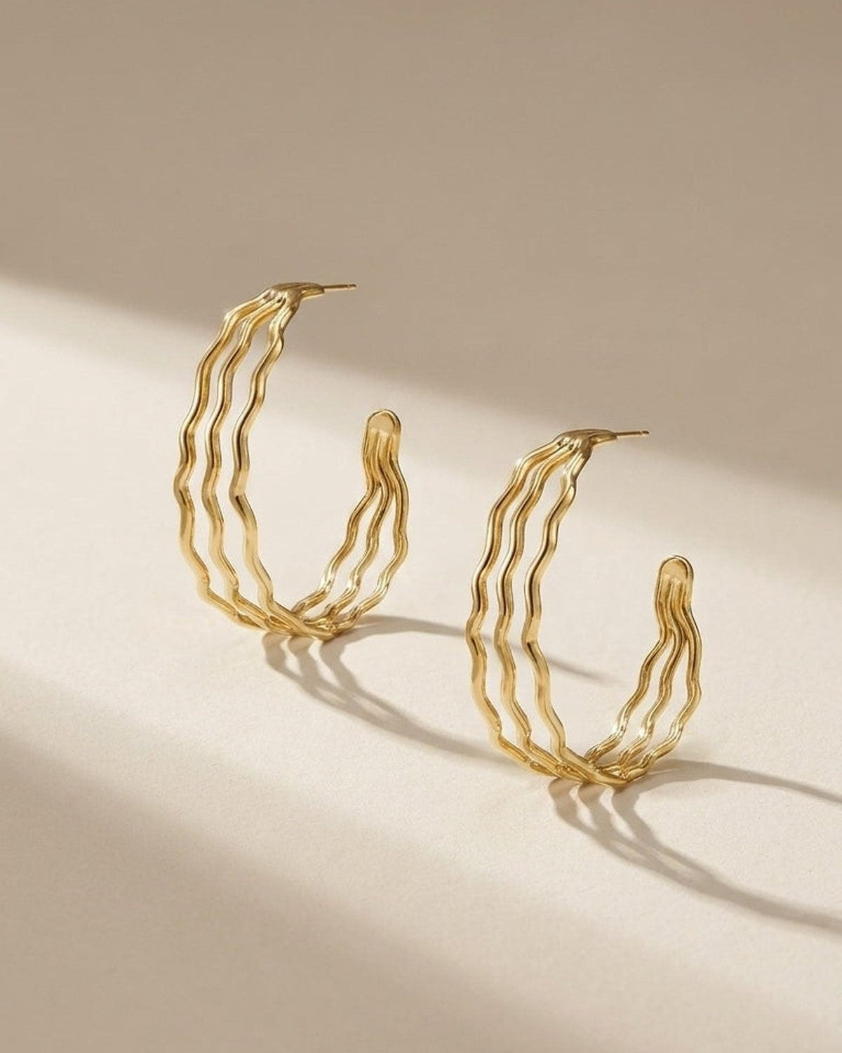 Two gold hoop earrings featuring a triple-band wavy design, displayed upright on a light surface.