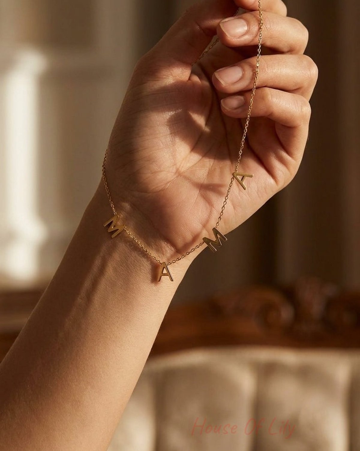 A delicate gold chain necklace spelling "MAMA" in hanging block letters, held in an open hand.