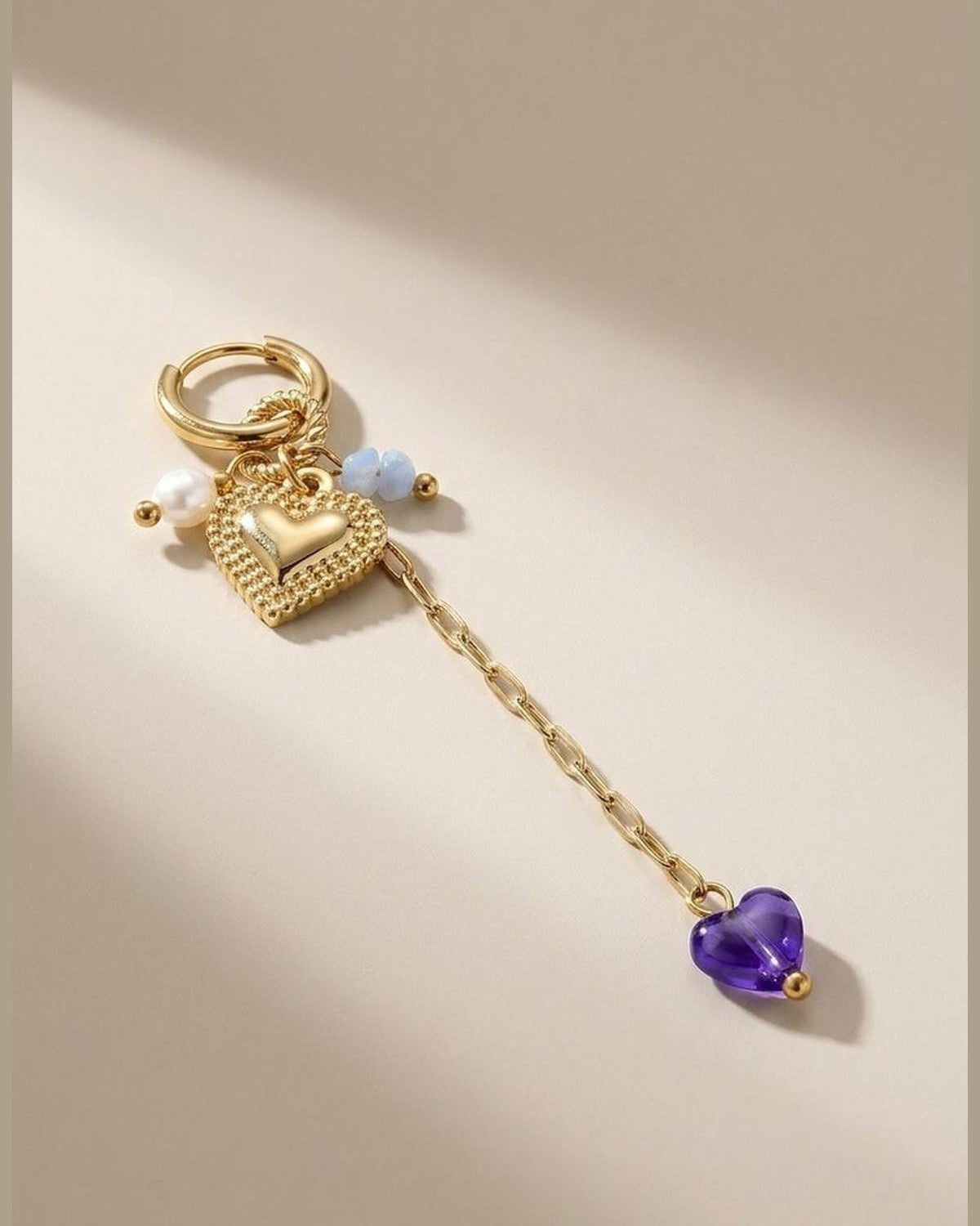 Gold hoop earring with a textured heart charm, pearl and blue beads, and a dangling chain with a purple heart.