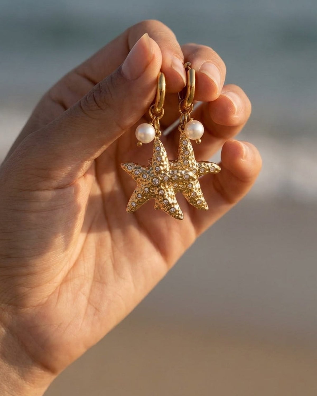 Gold hoop earrings with crystal-studded starfish charms and hanging white pearls, held against an ocean backdrop.