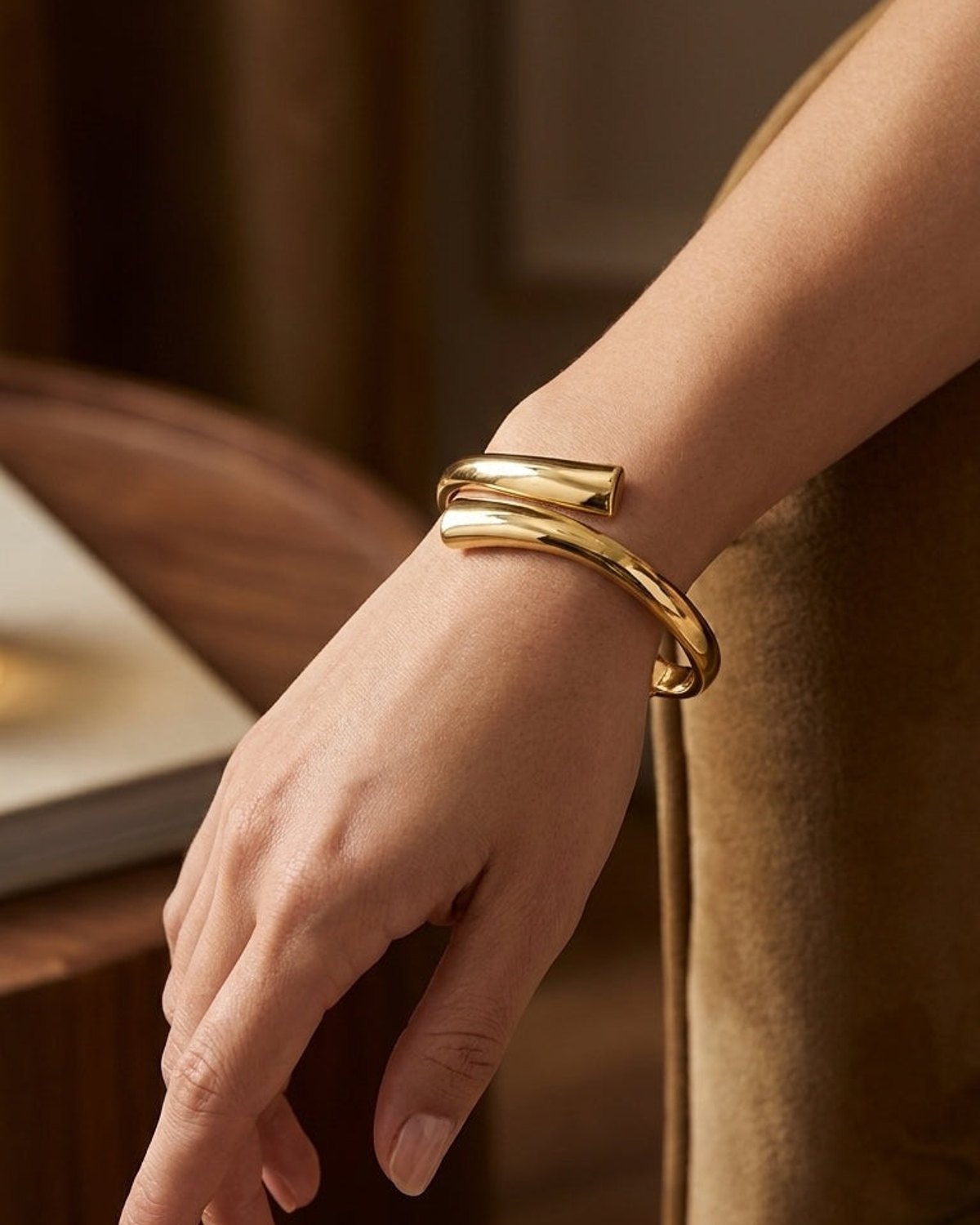 A shiny gold-plated cuff bracelet wraps smoothly around a wrist, featuring a sleek, modern open-ended design.