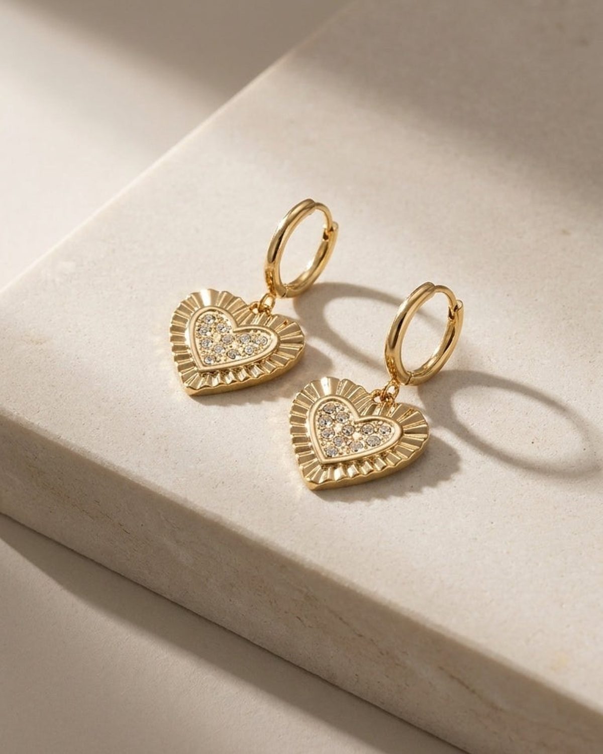 Gold hoop earrings featuring heart-shaped charms with crystal-paved centers and ridged edges on a stone surface.