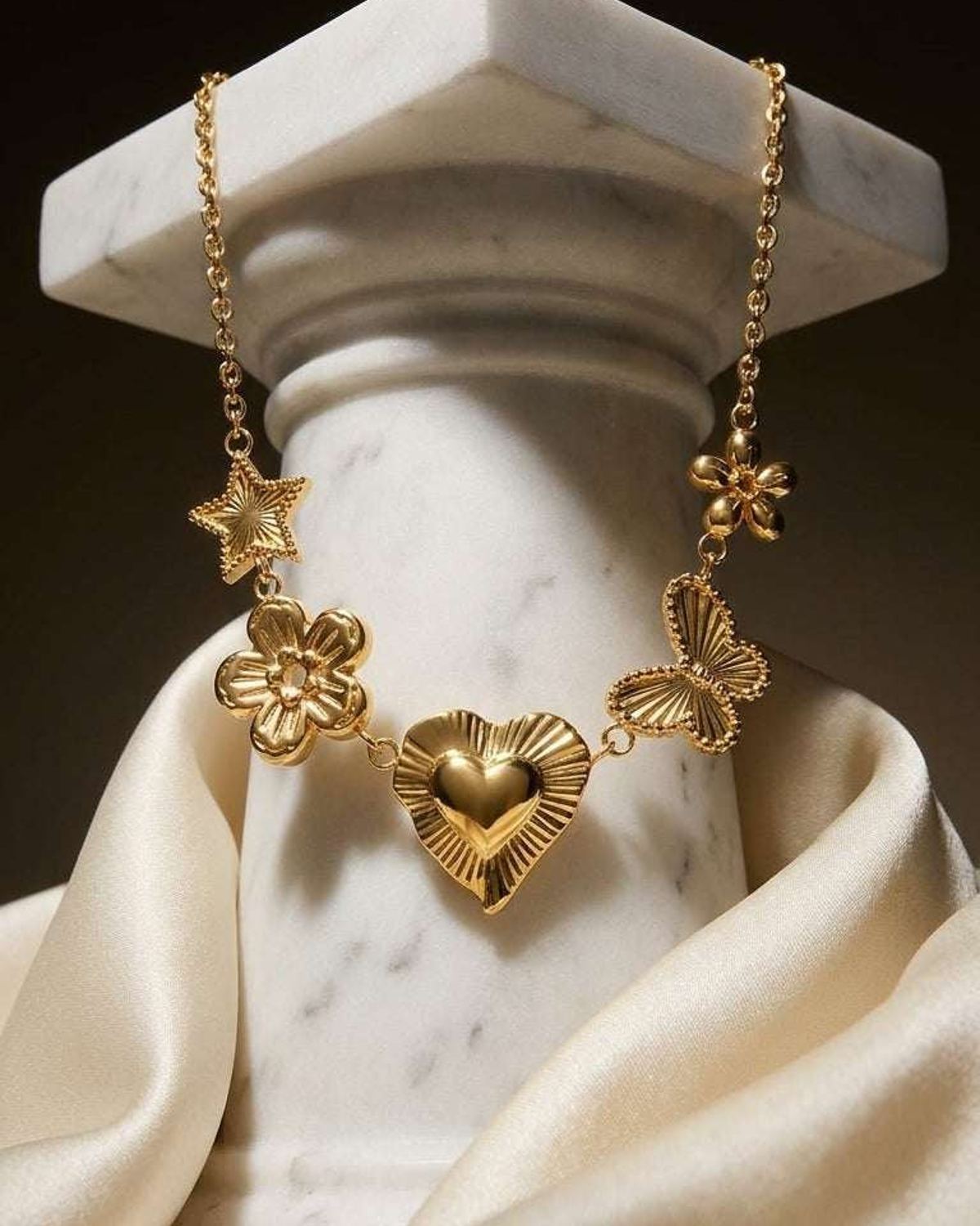 Gold chain necklace with charms of a star, flowers, heart, and butterfly draped over a white marble column.
