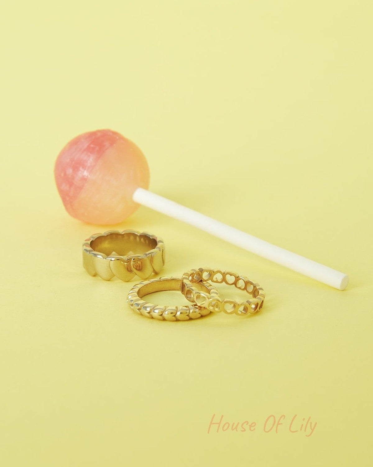 Three textured gold-tone rings arranged beside a pink and orange lollipop on a yellow background