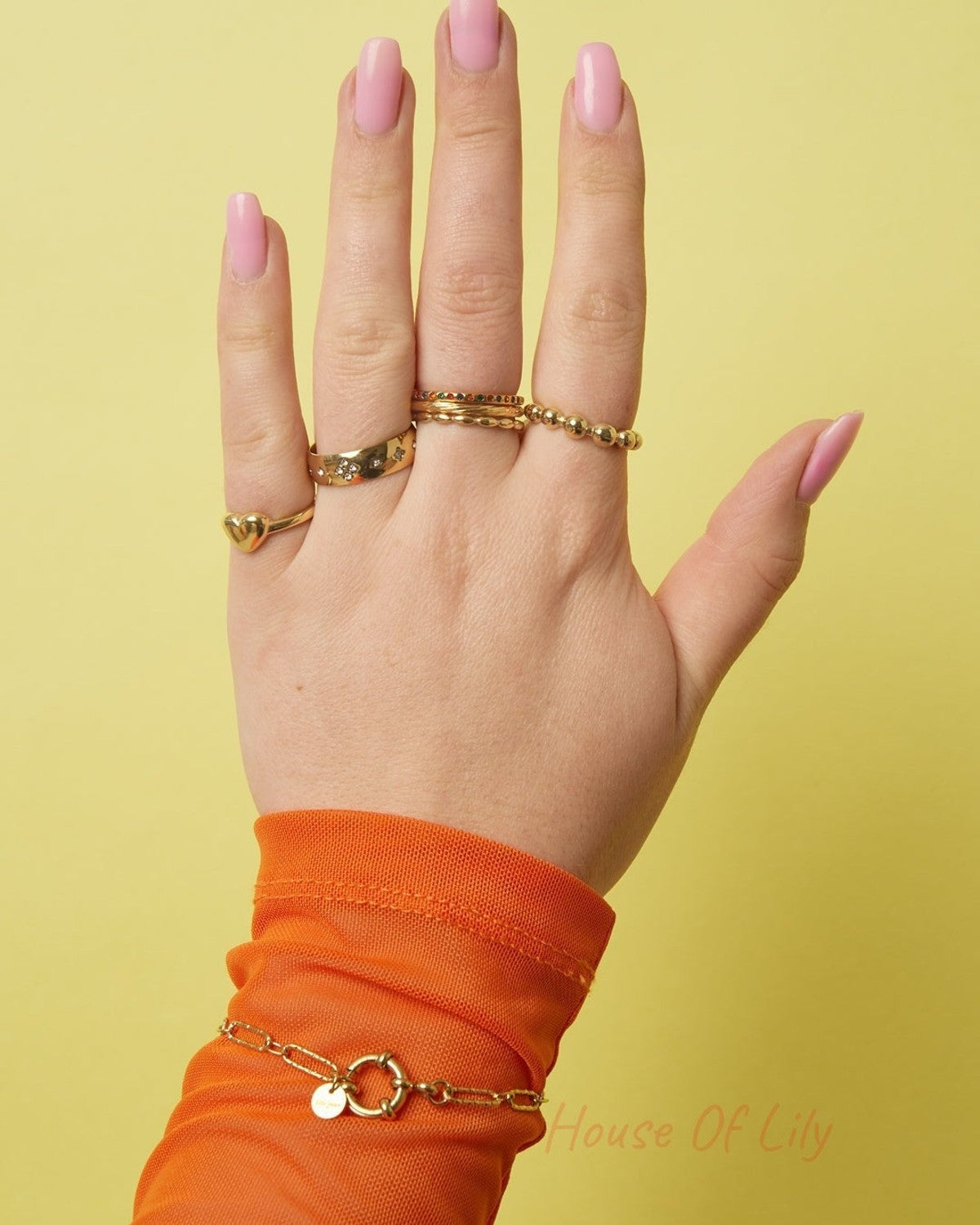 Hand wearing four gold rings including a beaded gold ring, displayed against a yellow background with pink nail polish and orange sleeve