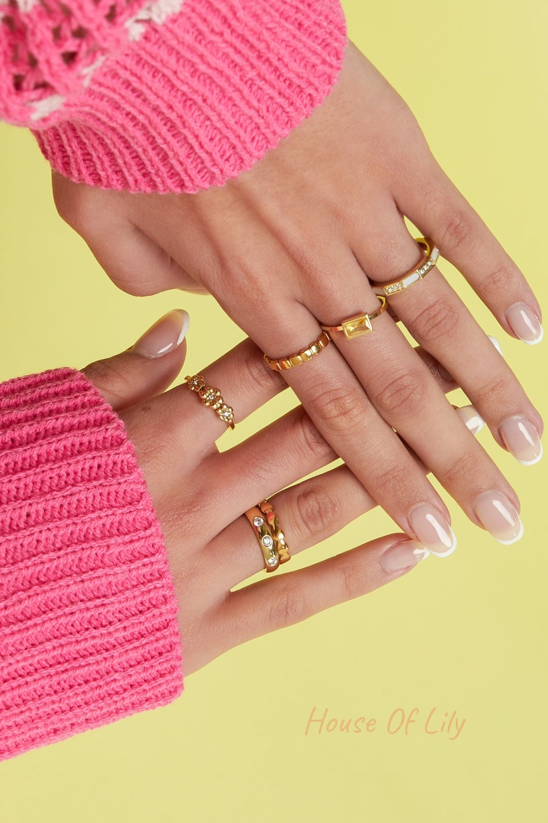 Close-up of hands wearing multiple gold rings including a rectangular yellow stone ring, with a pink knitted sweater and yellow background