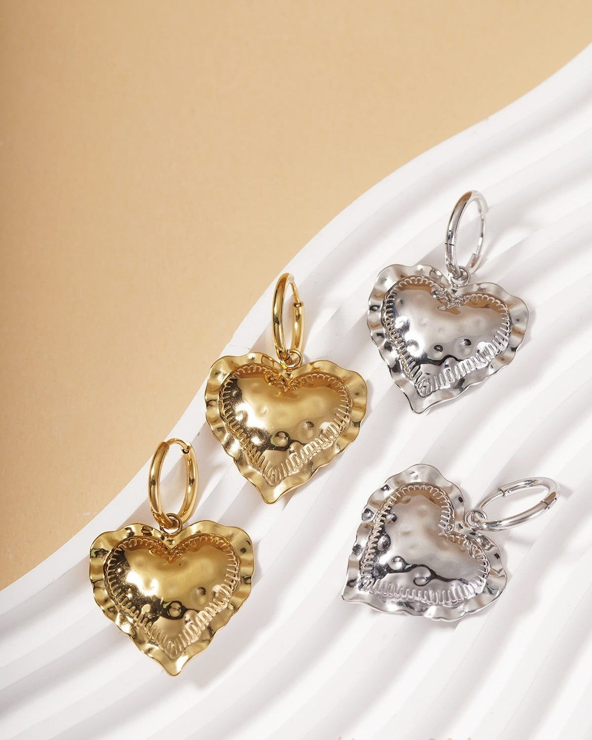 Heart Loop Drops: Gold and silver hoop earrings with textured, ruffled-edge heart charms on a white wavy display.