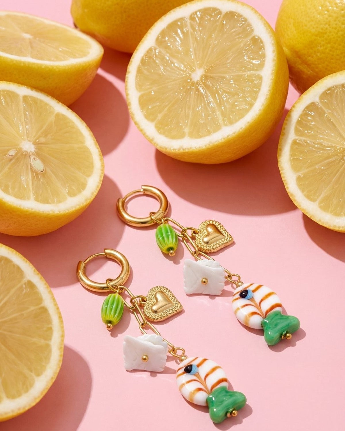 Gold-tone dangle earrings featuring charms of hearts, white squares, and fish, set against a pink background with lemons.