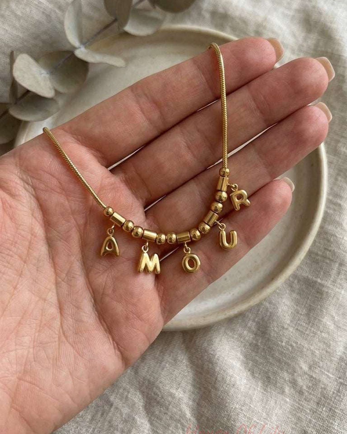 Gold Amour charm necklace held in a palm, featuring delicate letter charms and beads on a fine chain.
