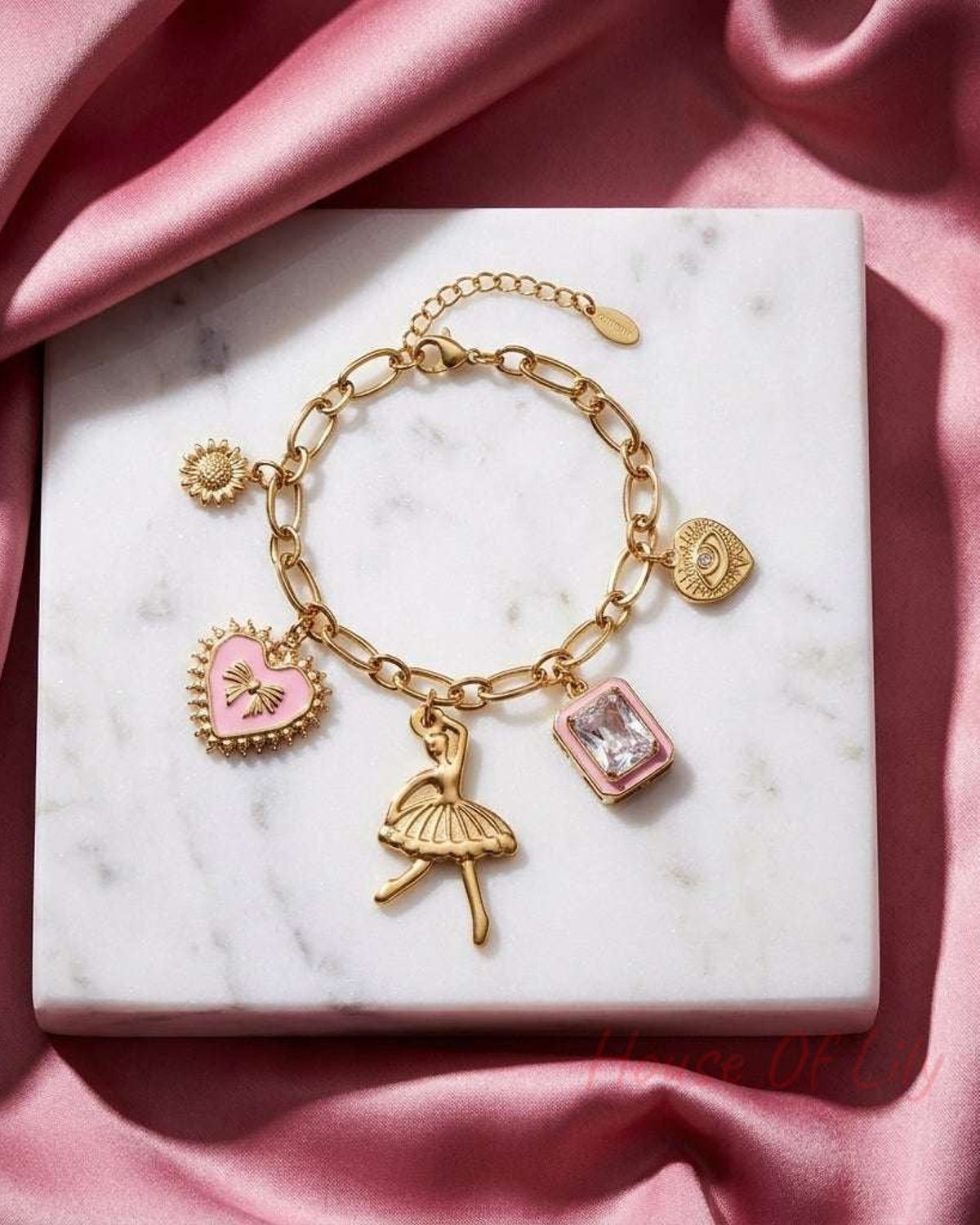 Gold chain charm bracelet with pink enamel heart, ballerina, crystal, and evil eye charms on a white marble base.