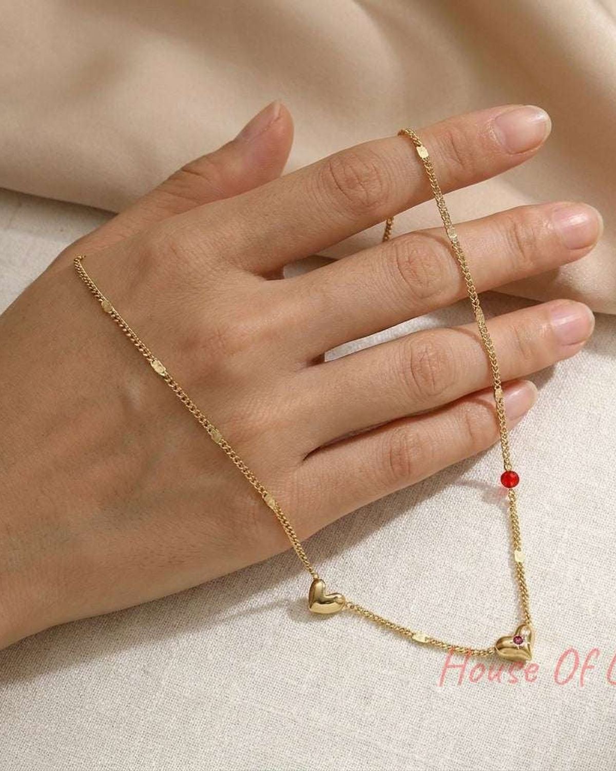 Gold chain necklace on a hand featuring small gold hearts, a red bead, and a tiny embedded pink stone.
