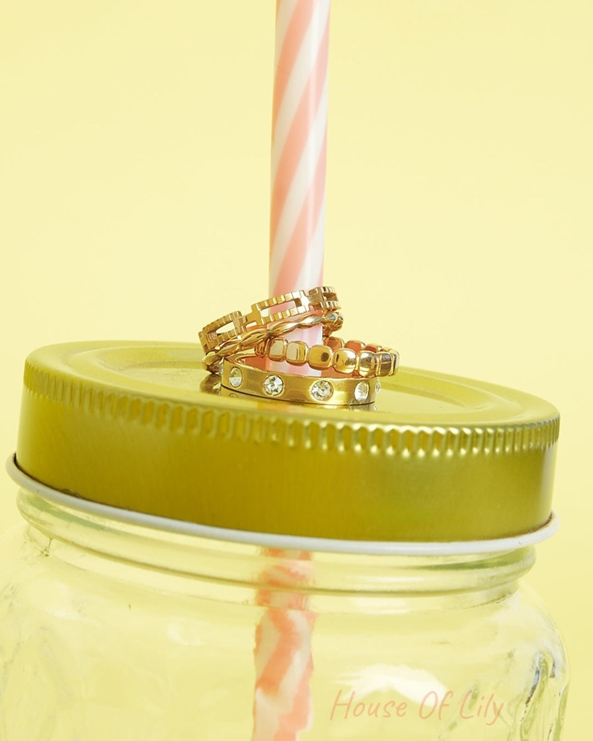Three gold rings stacked on a golden lid of a jar with a pink and white striped straw against a yellow background