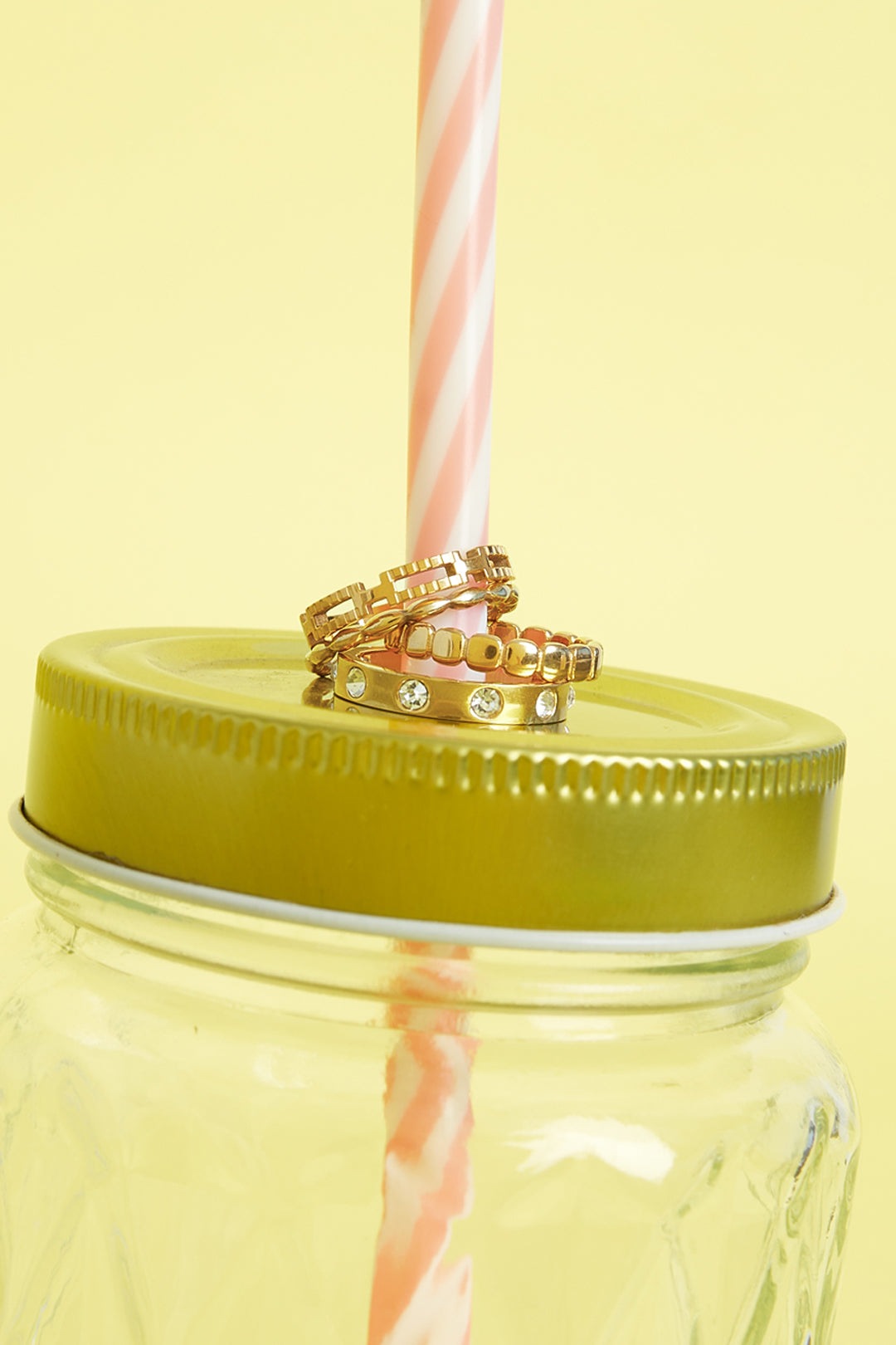 Three gold rings stacked on a golden lid of a jar with a pink and white striped straw against a yellow background