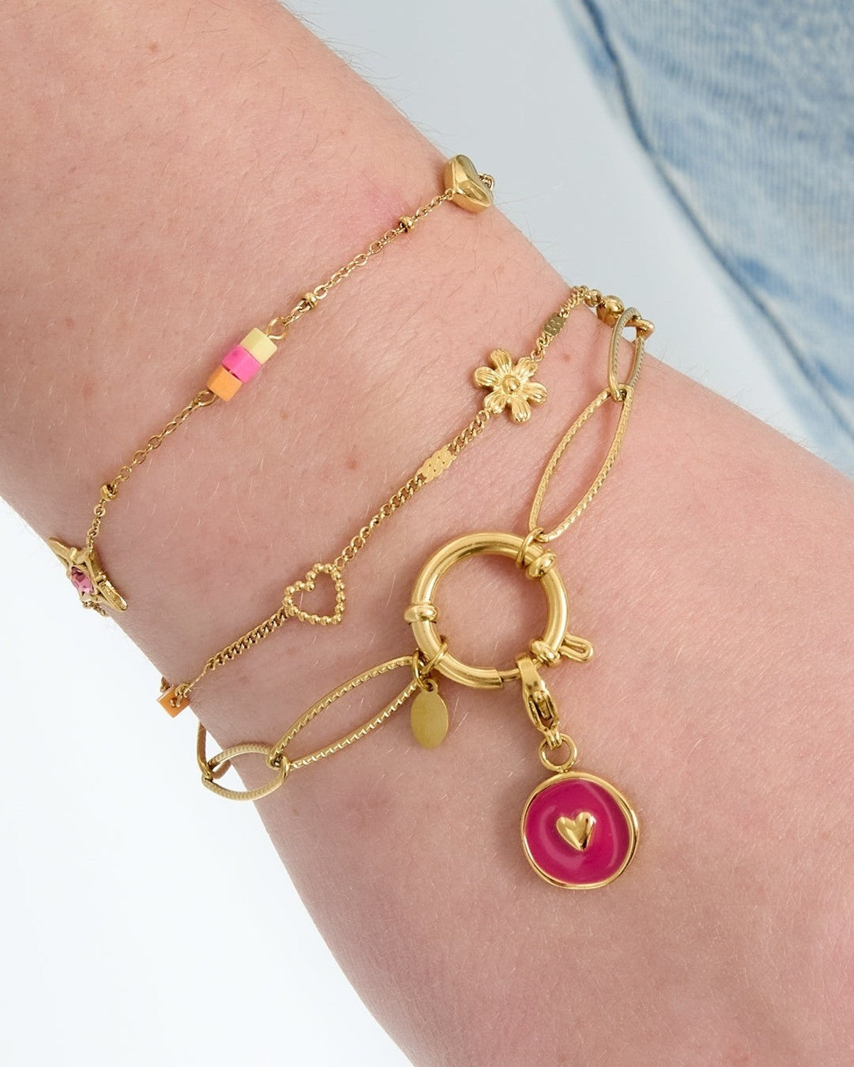 Close-up of a wrist adorned with layered gold bracelets, one featuring a gold link chain and toggle clasp with a pink charm, styled with denim