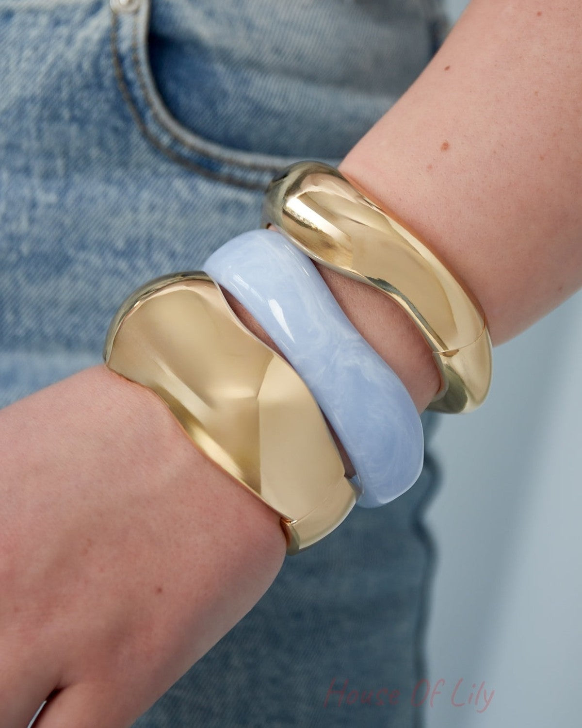 Close-up of a wrist wearing two golden sculpted bangles and one light blue resin bangle, styled with denim jeans