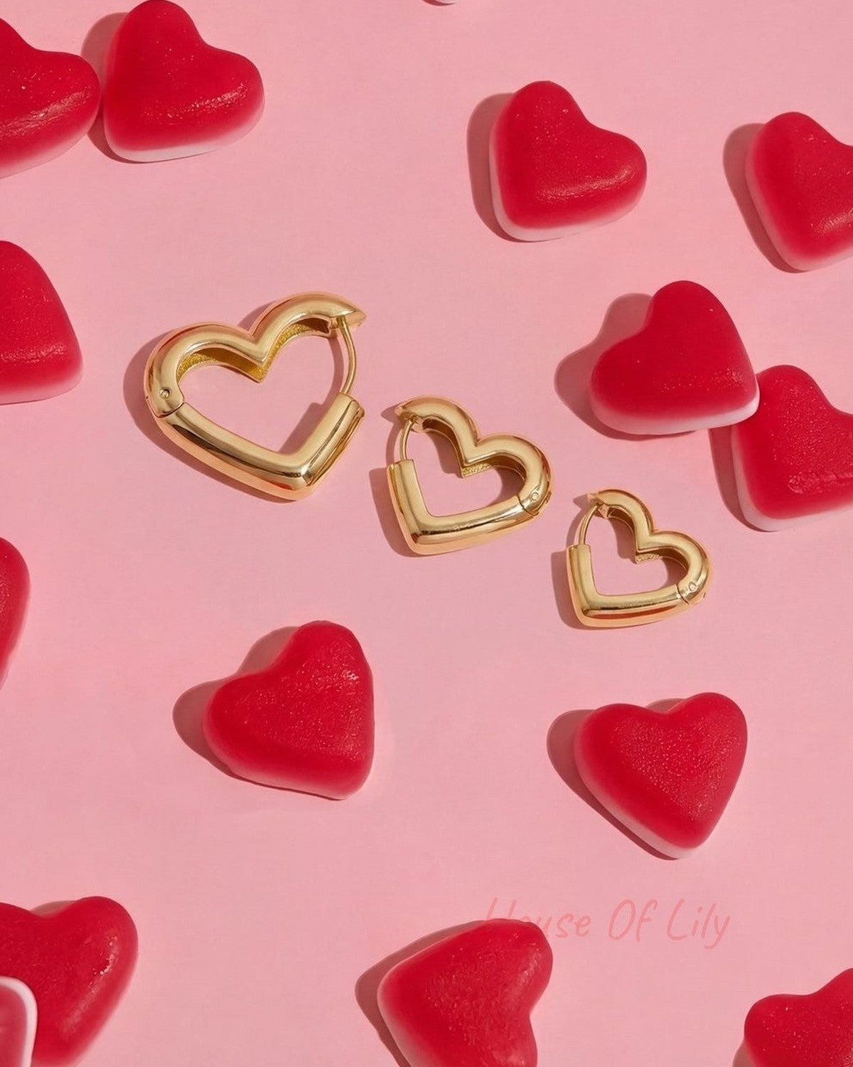 Three gold heart-shaped hoop earrings of varying sizes, scattered on a pink surface with red gummy heart candies.