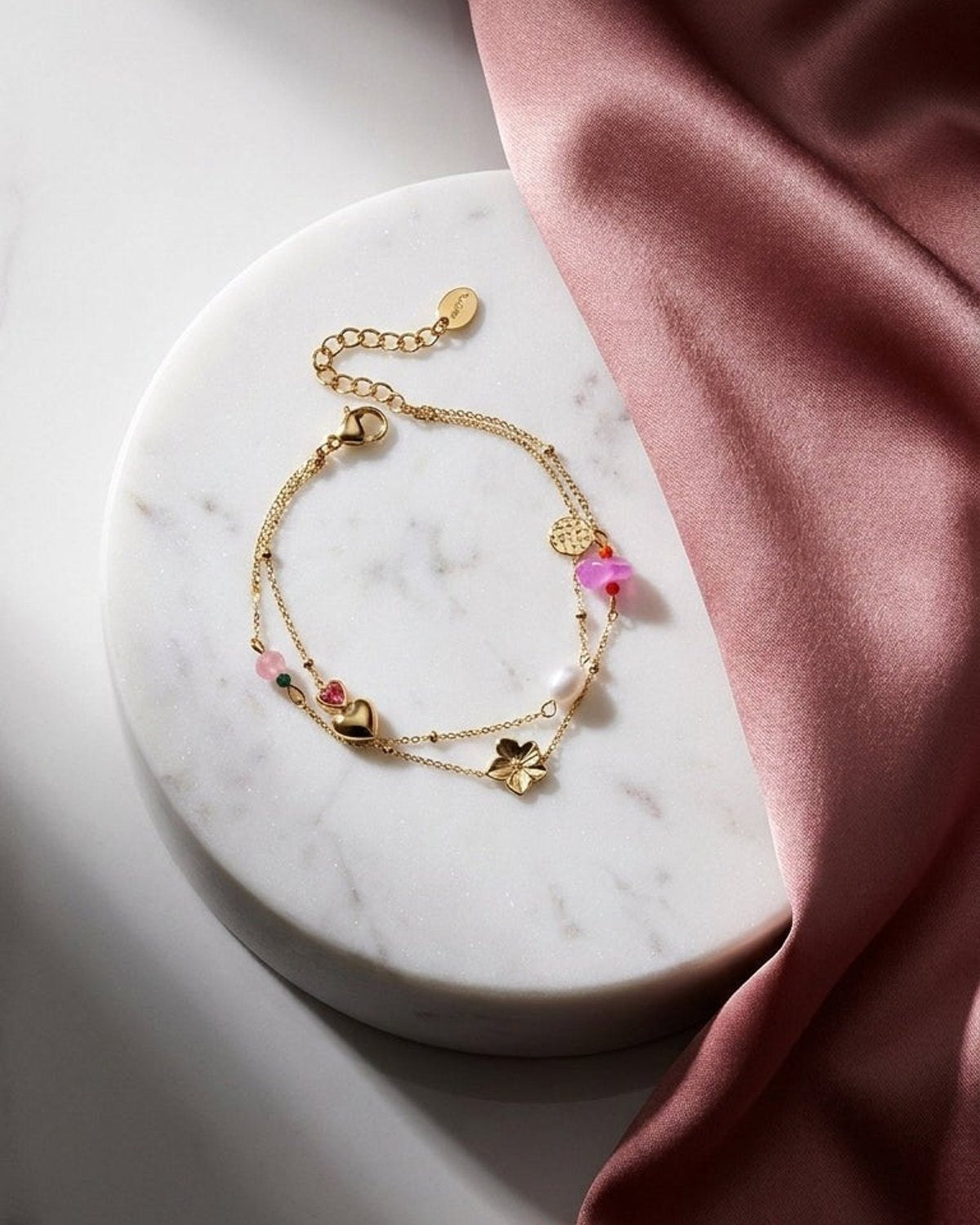 Gold charm bracelet stack with a heart, flower, pearl, and pink stones on a marble surface with pink silk.