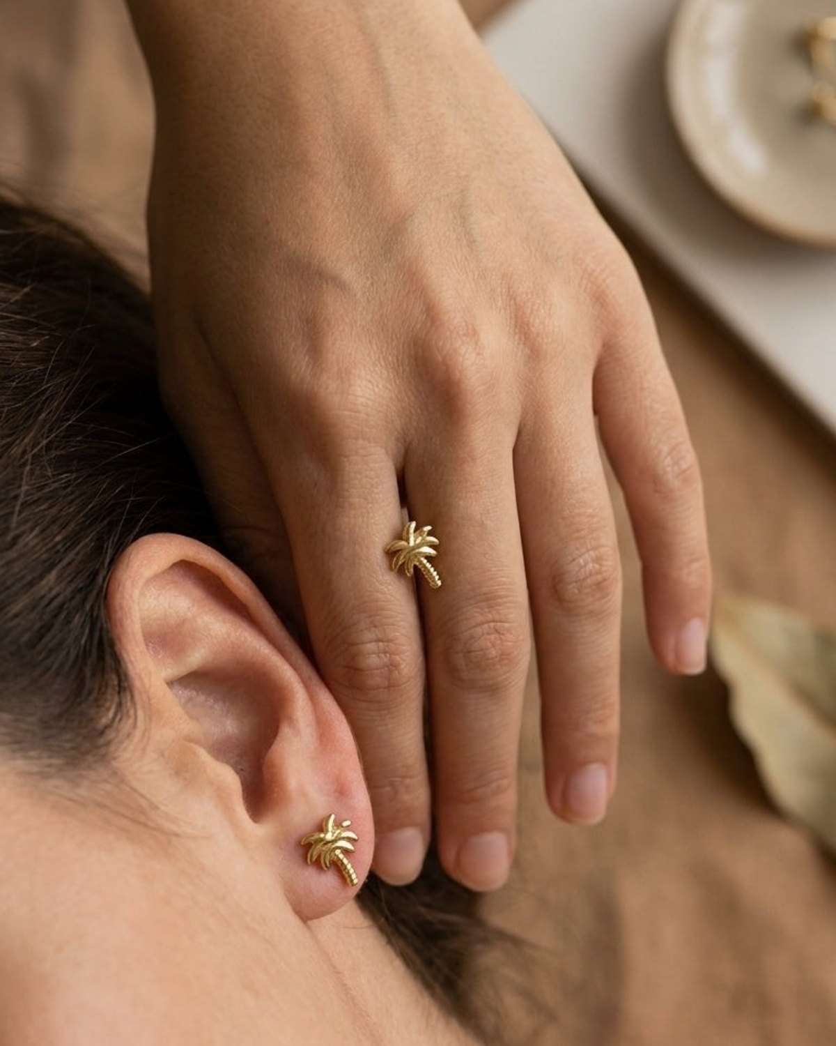 Gold palm tree stud earring and matching ring on a model against a neutral background.