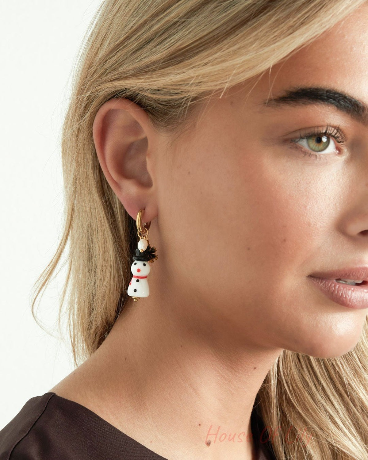 Close-up of a woman wearing a gold-tone hoop drop earring with a dangling white resin snowman charm, black hat, red scarf, gold snowflake, and faux pearl.