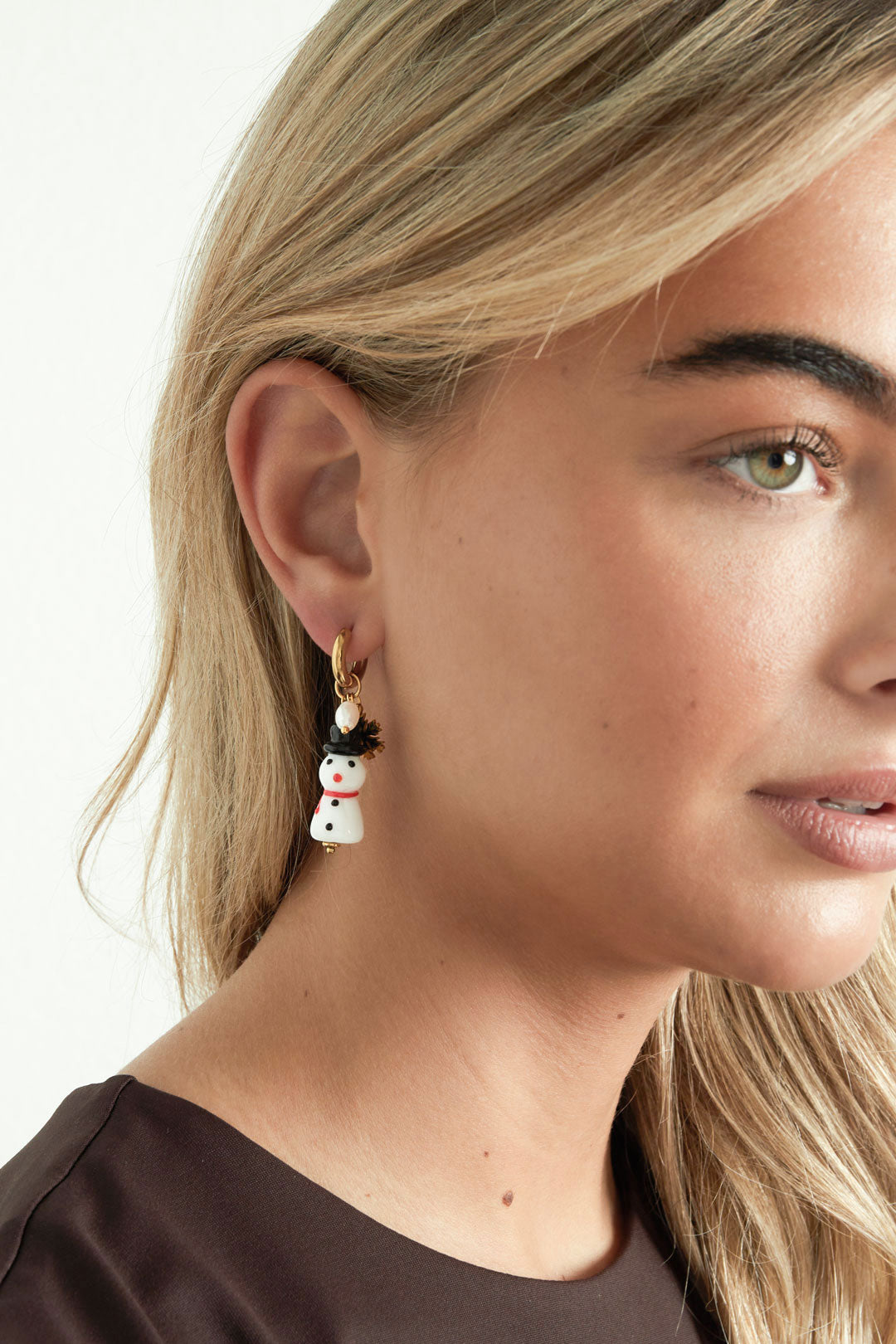 Close-up of a woman wearing a gold-tone hoop drop earring with a dangling white resin snowman charm, black hat, red scarf, gold snowflake, and faux pearl.