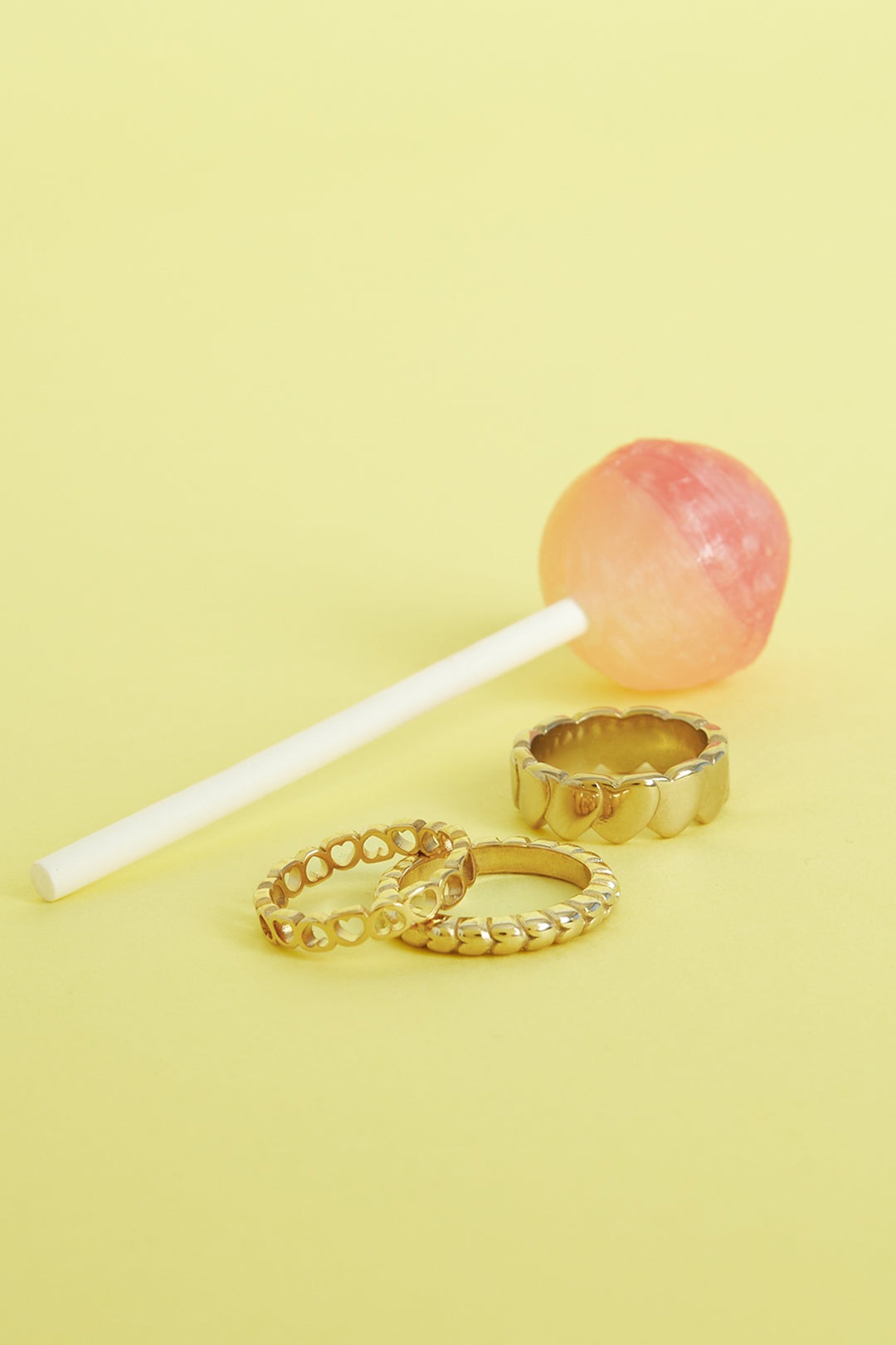 Three gold-tone rings with heart patterns placed on yellow background beside a pink and orange lollipop