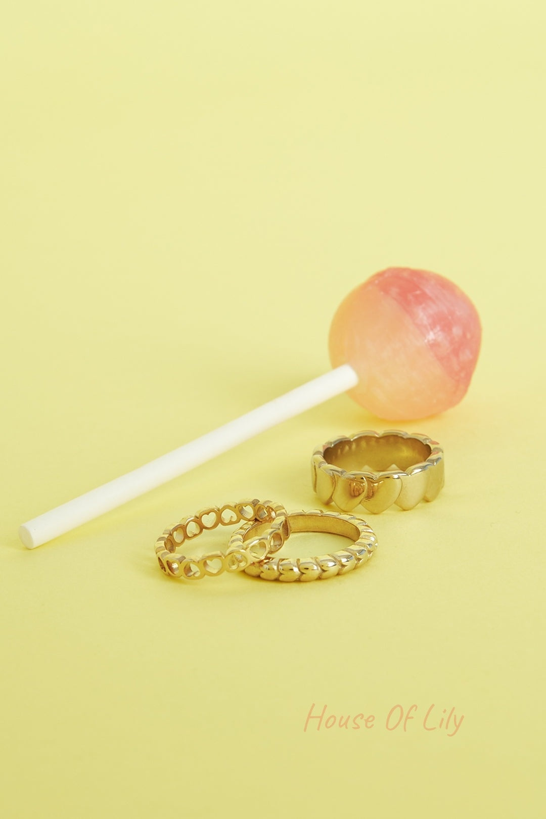 Three gold-tone rings with heart patterns placed on yellow background beside a pink and orange lollipop
