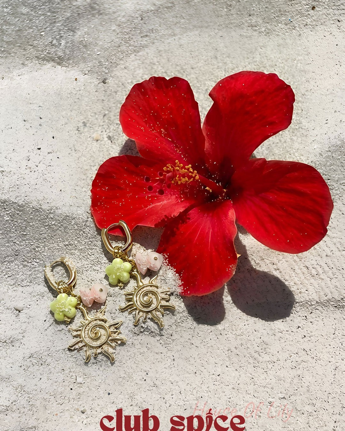 Gold-tone sun charm drop earrings displayed next to a red hibiscus flower on a textured stone surface with Club Spice branding