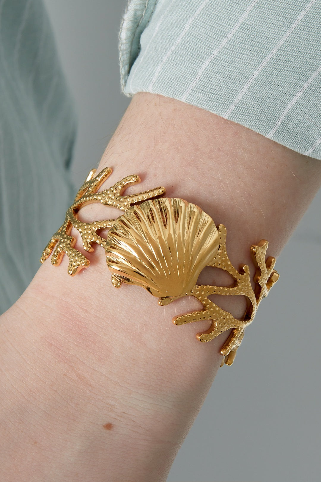 Close-up of golden seashell cuff bracelet worn on wrist with striped light blue shirt