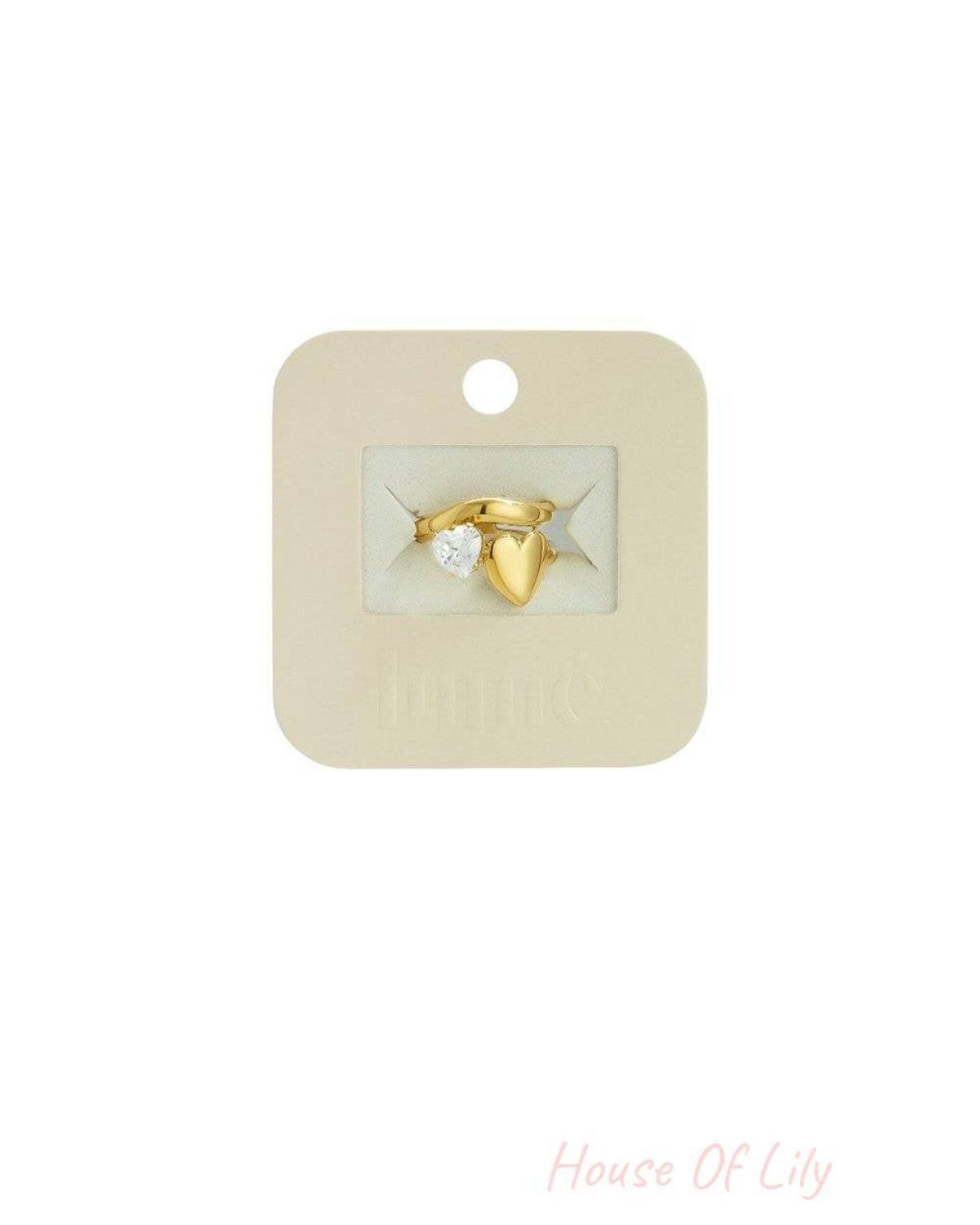 Gold-tone ring featuring a clear heart-shaped gem and a solid polished heart charm on a branded display card.