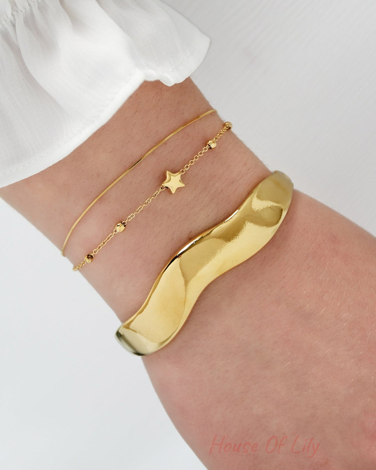 Close-up view of wrist wearing two gold bracelets; one delicate star charm bracelet paired with a smooth, wavy polished gold bangle and white sleeve.